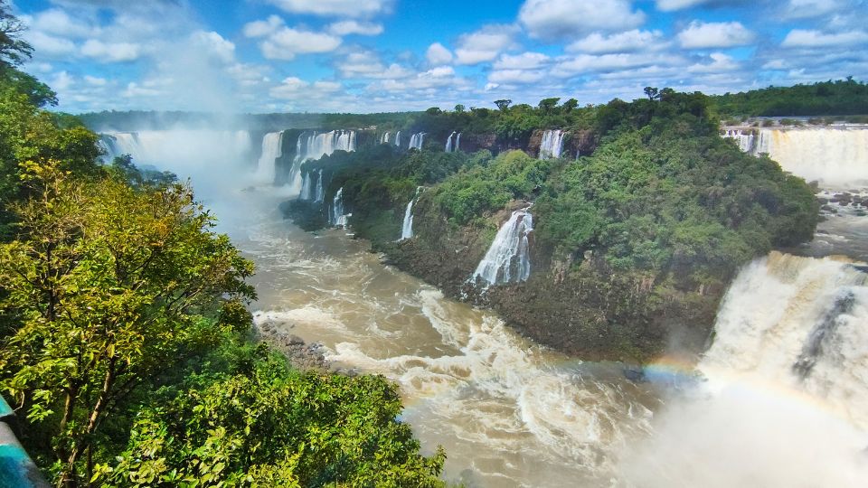 As Cataratas do Rio Iguaçu, uma das Sete Maravilhas da Natureza. a Foto mostra uma sequencia de quedas de água, o verde da mata preservada e o bonito céu azul. 