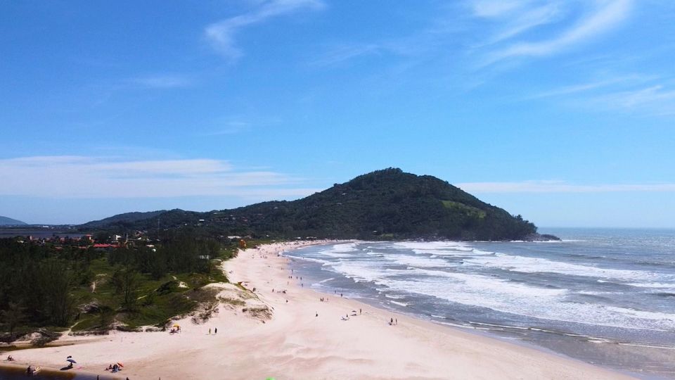 A linda Praia da Ferrugem, com suas areias fofas, brancas avermelhadas, mar com espuma branca e o azul refletindo o céu e paisagem de mata preservada.