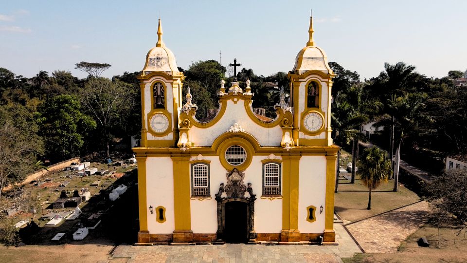 Igreja Matriz de Santo Antônio em Tiradentes. Uma igreja branca com detalhes em amarelo, com duas torres e um frontão bem adornado, do lado esquerdo existe um cemitério