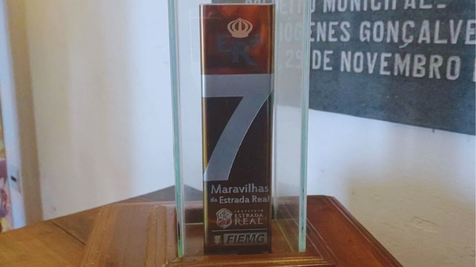 Um troféu de três faces, dourado com um numeral 7 estampado em cada face abaixo do numeral Maravilhas da Estrada Real